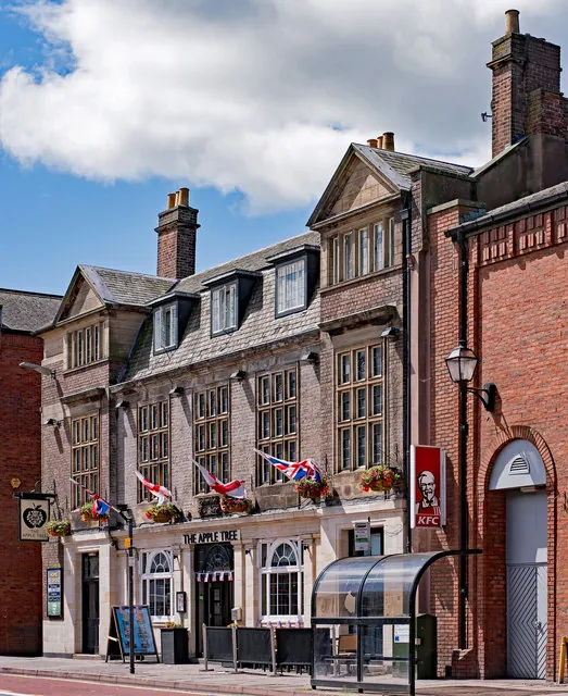 Three-storey stone building with a slate roof, dormer windows and a pedimented gable, its ground floor lettered The Apple Tree beneath a row of Union flags and hanging baskets, with a hanging apple sign at the left corner and a red brick KFC building and bus shelter to the right