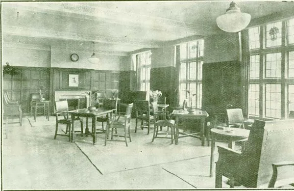 Old printed photograph of a spacious panelled room with tall leaded windows, low tables with wooden armchairs, a fireplace with a picture above it, a clock on the wall and vases of flowers