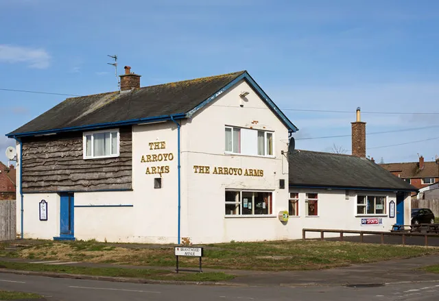 Two-storey whitewashed pub with blue trim, part clad in dark weatherboarding, The Arroyo Arms lettered in gold on the front and gable, and a lower single-storey wing to the right, on a grassy corner by a Brantwood Avenue street sign