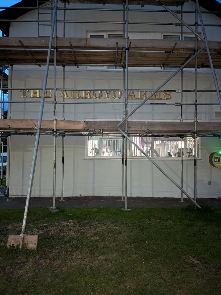 Gable end of a whitewashed brick pub at dusk, with The Arroyo Arms in gold letters across it, scaffolding in front, lit windows on the ground floor and grass in the foreground