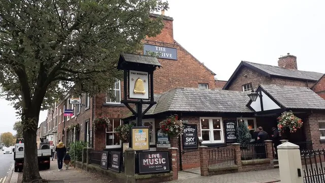 Red brick pub with a gable lettered The Beehive and a long single-storey slate-roofed front extension with white windows and hanging flower baskets. A post sign showing a golden beehive stands on the pavement beside a Live Music board, with a large tree and a few people on the street to the left.