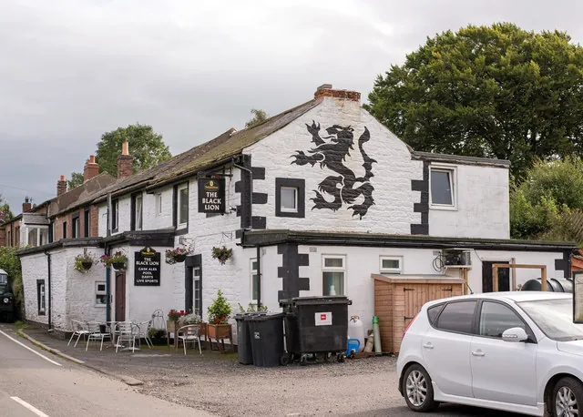 Long low whitewashed pub with black painted corner stones and window surrounds, a large black lion rampant painted on the gable, hanging signs reading The Black Lion, tables and flower baskets outside and a car parked in front