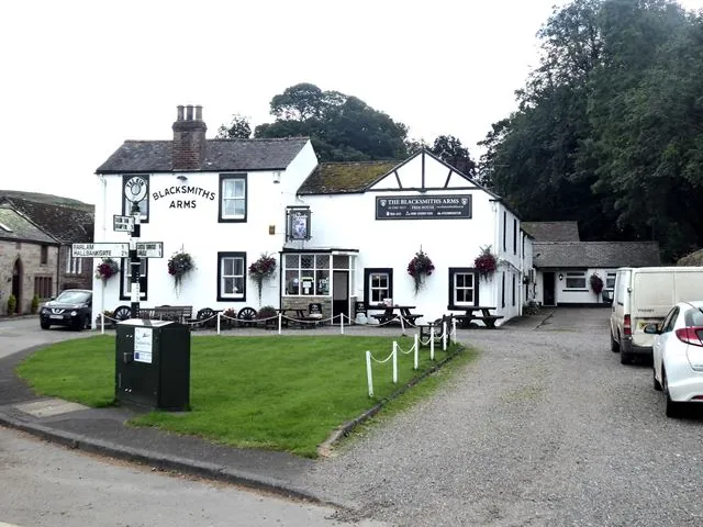 Whitewashed village pub with a slate roof and a mock Tudor gable, Blacksmiths Arms painted on the end wall and a sign reading The Blacksmiths Arms Free House, with hanging baskets, picnic benches and old cart wheels in front, a small village green and fingerpost signs at the corner