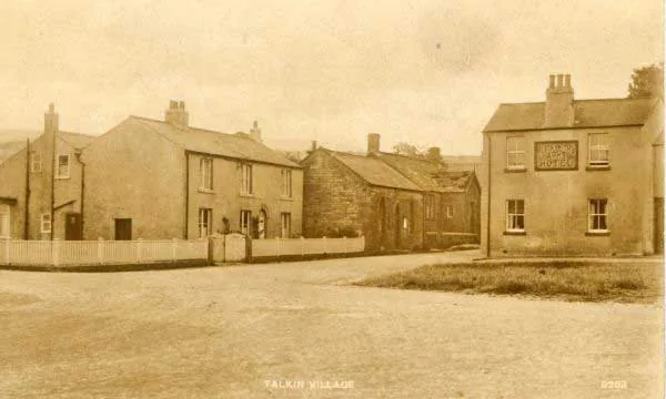 Old sepia postcard of a village green with low cottages and a chapel, and on the right a square two-storey rendered building with a signboard for the hotel, captioned Talkin Village