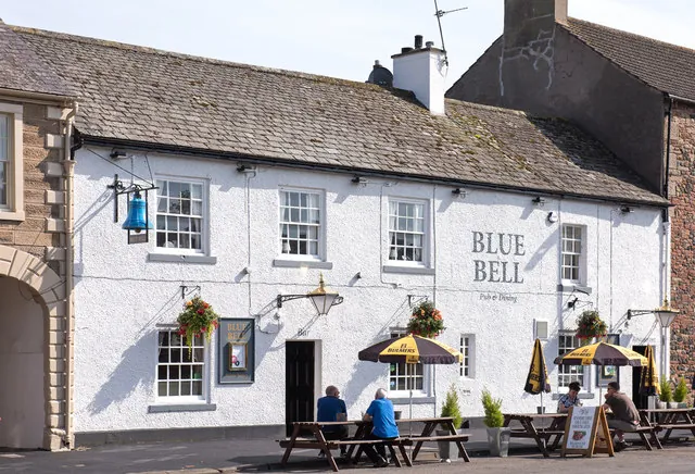 Whitewashed two-storey pub with a grey slate roof and white sash windows, the name Blue Bell in grey letters on the front and a blue bell hanging from a bracket at one end. Picnic benches and parasols stand outside with people sitting at them, beside a sandstone arch on the left.