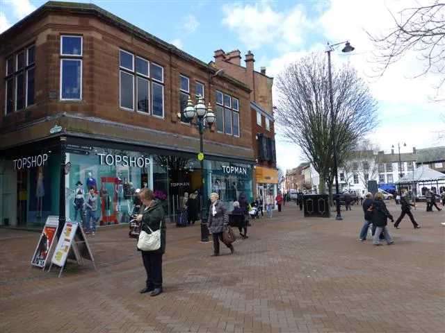 Two-storey red sandstone corner building with large upper windows above glazed shopfronts lettered Topshop and Topman, on a busy paved pedestrian street with shoppers, lamp posts and bare trees