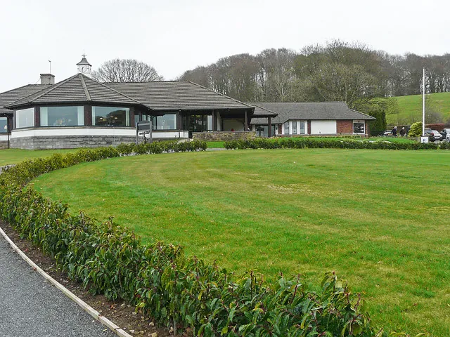 Low single-storey clubhouse with a wide hipped roof, a small cupola and long windows, seen across a curving clipped hedge and a sweep of mown grass with wooded hills behind