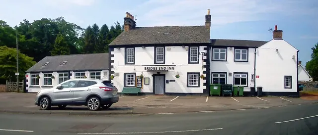 Long white rendered pub with a grey slate roof, black window surrounds and quoins, and a sign reading Bridge End Inn above the central door. Lower white wings extend either side, wheelie bins stand against the wall and a silver car is parked on the forecourt by the road.