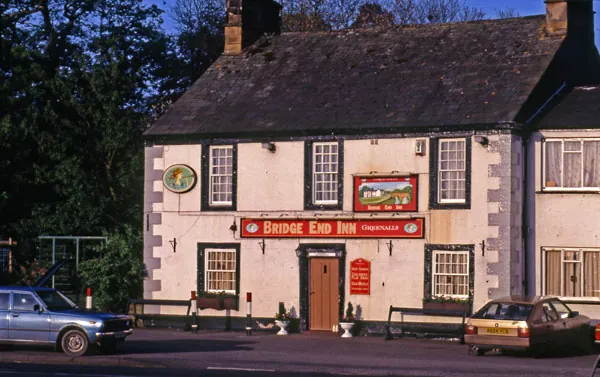Colour photograph of a two-storey whitewashed inn with painted corner quoins, a long red sign reading Bridge End Inn Greenalls, a painted picture board between the upper windows, and cars parked on the road in front