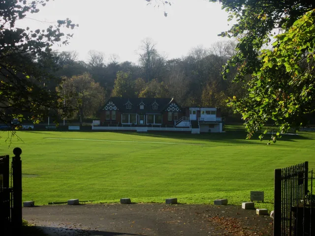 Mock Tudor cricket pavilion with black and white gables and a white scorebox alongside, seen in the distance across a broad sunlit cricket field through open iron gates, with autumn woodland behind