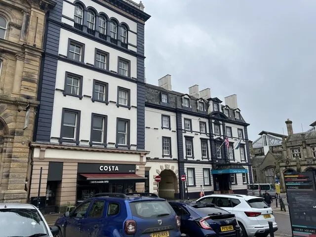 Three and four-storey white painted buildings with dark grey window surrounds and dormers, a Costa shop at street level and a teal canopy over the hotel entrance, with flags above and a row of parked cars in front