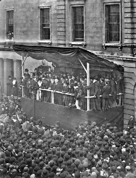 Black and white photograph looking down on a dense crowd in bowler and top hats around a draped wooden platform against the stone front of a large classical building, with men in top hats packed onto the platform