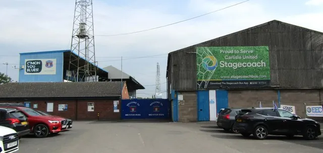 Football ground entrance with blue gates reading Welcome to Brunton Park, a blue stand behind with a sign reading C'mon You Blues, a floodlight pylon, and a large grey shed covered by a green Stagecoach banner, with cars parked in the foreground
