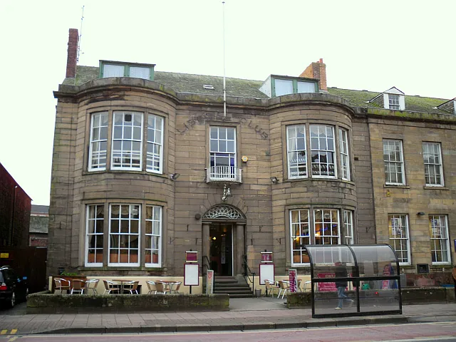 Three-storey sandstone building with two full height curved bay windows flanking a central doorway with a fanlight, a small first-floor balcony, faint carved lettering across the upper front and dormer windows in the roof, with tables and chairs on a raised terrace and a glass bus shelter on the pavement