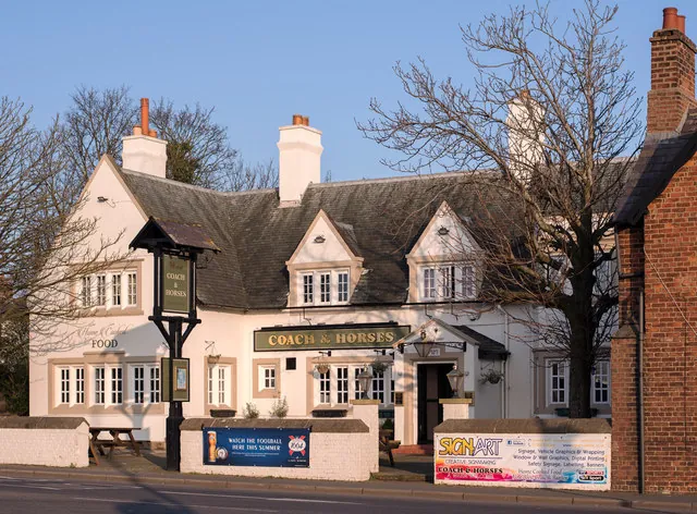 Long whitewashed pub with a grey slate roof, two gabled dormers and tall white chimneys, a dark sign reading Coach & Horses above the entrance and a tall post sign at the front, lit by low sunshine behind a low white wall with bare trees around it