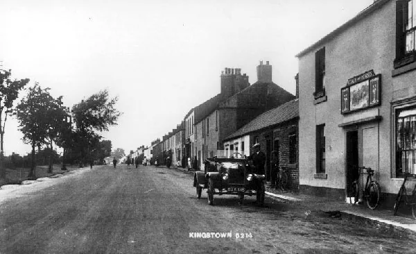Old postcard view along a wide village road lined with low cottages, an early open motor car standing outside a pub on the right with a picture sign above the door and bicycles propped against the wall, captioned Kingstown