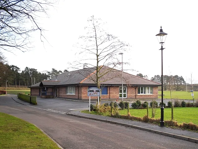 Single-storey red brick clubhouse with a hipped tiled roof and a row of white-framed windows, set back across a tarmac drive with a lamp post, a young tree and a Creighton sign in the foreground and pitches beyond