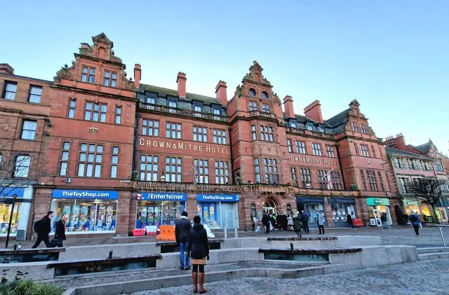 Long four-storey red sandstone frontage with ornate Dutch gables and the words Crown and Mitre Hotel along the upper floor, shops on the ground floor and people crossing a paved square in front