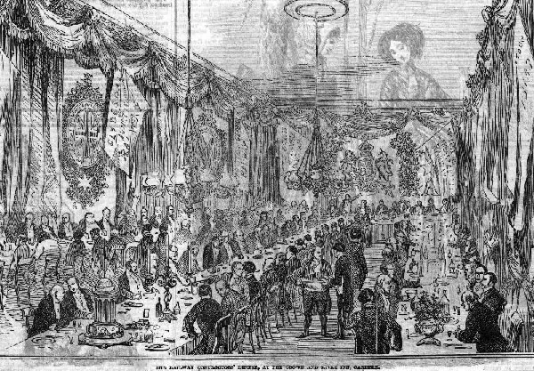 Engraving of a long banqueting room hung with drapery, flags and coats of arms, with chandeliers above two long tables lined with men in dark coats at dinner and waiters moving between them