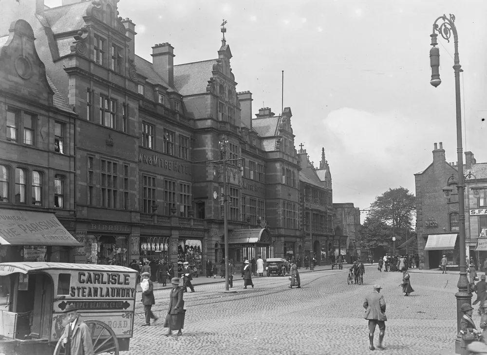 Black and white photograph of the long red sandstone frontage of the Crown and Mitre Hotel, with Dutch gables, its name along the first floor and a glazed canopy over the entrance, a horse-drawn laundry cart lettered Carlisle Steam Laundry in the foreground and people crossing the cobbled street