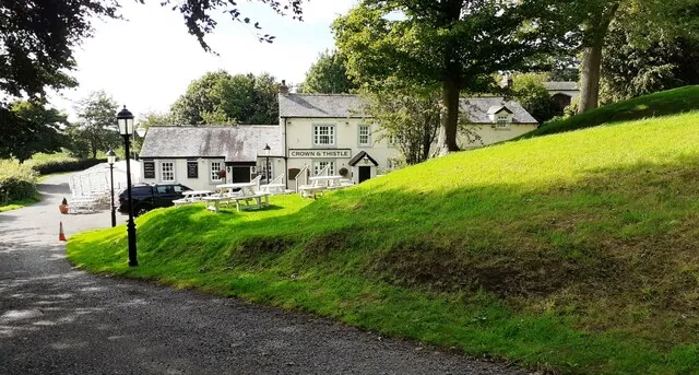 Low whitewashed pub with a slate roof and a sign reading Crown and Thistle over the door, picnic benches and a parked car in front, seen past a steep grassy bank and a lane lined with black lamp posts under mature trees