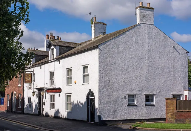 Two-storey whitewashed pub with a slate roof and small dormers, a red fascia reading Crown & Thistle and a hanging pictorial sign over the door, and a broad blank white gable end facing the street