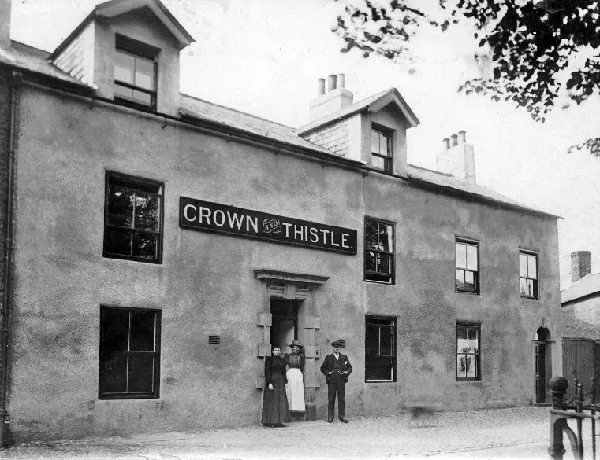 Old photograph of a long two-storey rendered building with dormers and a large board reading Crown and Thistle above the doorway, where two women, one in a white apron, and a man in a cap stand
