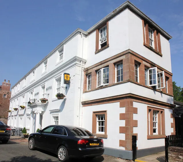 Three-storey white painted hotel with red sandstone window surrounds and corner stones, a projecting bay at the corner, flower baskets along the front and a car parked outside