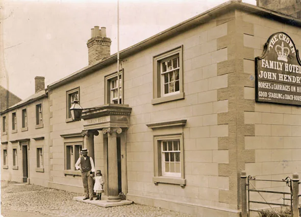 Old sepia photograph of a long two-storey stone inn with a columned porch and a lantern, a man in a waistcoat and a small girl at the door, and a board reading The Crown Family Hotel, John Hendry, horses and carriages on hire, good stabling