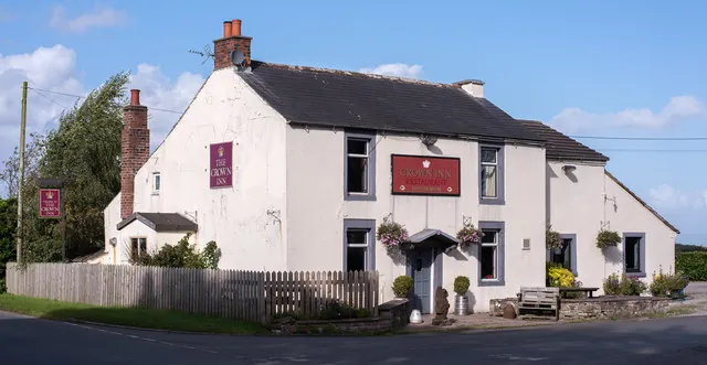 Whitewashed two-storey country pub with a slate roof and brick chimneys, maroon signs reading The Crown Inn and Crown Inn Restaurant, hanging baskets around the doorway and a wooden fence along the roadside