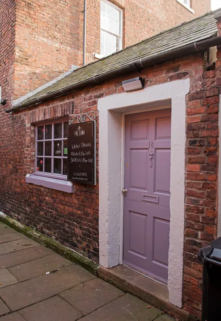 Small single-storey red brick building with a slate roof tucked against a taller brick building, with a lilac painted panelled door numbered 18 in a white surround. A black board reading The Lane, with chalked winter opening hours, hangs beside a small multi-paned window above a flagstone pavement.