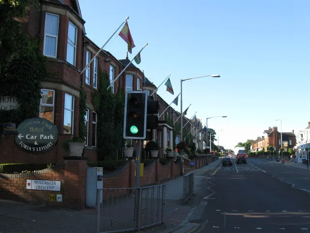 Row of tall red brick Victorian houses with bay windows and a line of flagpoles flying flags, seen in shadow along a wide main road. A round sign reading Cumbria Park Hotel, Car Park and Toria's Leisure stands by the low front wall next to a Mulcaster Crescent street sign and a traffic light showing green.