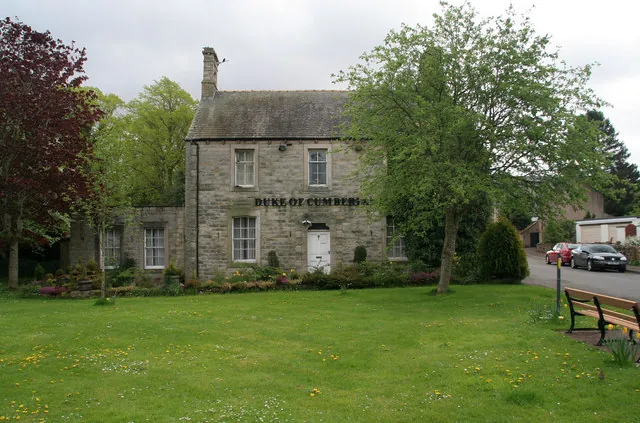 Two-storey grey stone pub with a slate roof and tall chimney, a white door and the words Duke of Cumberland painted across the front, partly hidden by a leafy tree. A lower stone wing sits to the left and a wide village green with a wooden bench fills the foreground.