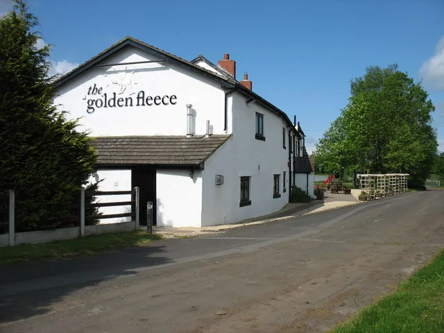 Long whitewashed inn with a slate roof and red brick chimneys, its gable end painted with the words the golden fleece, beside a country lane with trees on either side