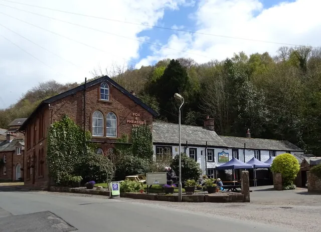 Red sandstone building with a tall gable and round-arched windows, lettered Fox and Pheasant Inn on the gable, joined to a long whitewashed cottage range with blue gazebos and outdoor tables, set against a steep wooded hillside
