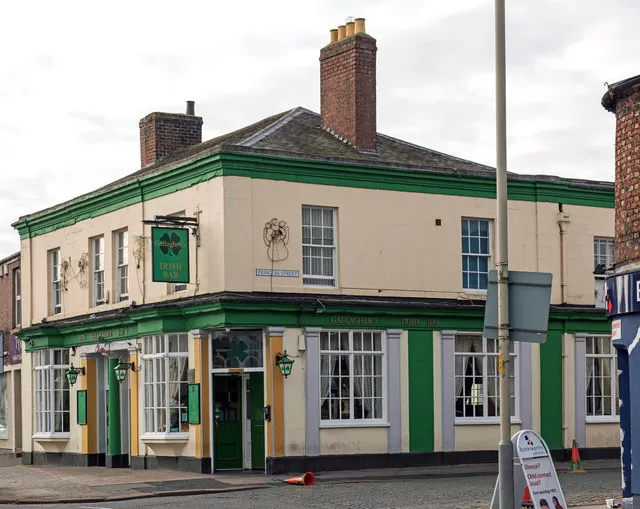 Two-storey cream painted corner pub with a slate roof, tall brick chimney and green painted eaves, and a green and gold ground-floor frontage with a green door, shamrock decoration and a hanging sign reading Gallagher's Irish Bar