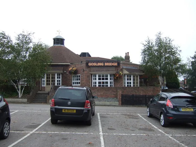 Low red brick pub with a fascia sign reading Gosling Bridge, white framed windows and hanging baskets, and a round tiled roof topped by a small white lantern and weathervane at one end. Trees stand either side and parked cars fill the tarmac car park in the foreground.
