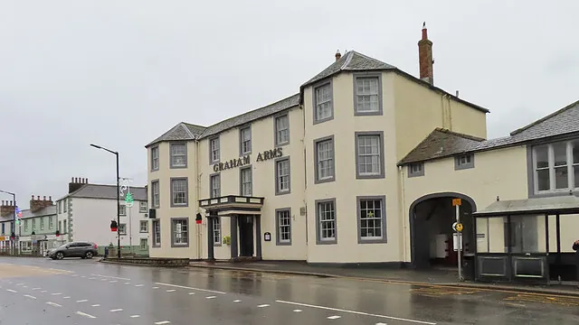 Three-storey cream rendered coaching inn with grey window surrounds and a slate roof, large letters reading Graham Arms across the front, a small columned porch over the door and an arched carriage entrance at the right end, beside a wet street on a grey day