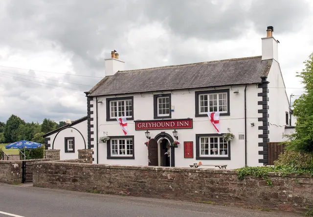 Two-storey whitewashed inn with a slate roof, black window surrounds and black corner quoins, a red sign reading Greyhound Inn above an arched doorway, and St George flags hung from the upstairs windows, behind a low red sandstone wall