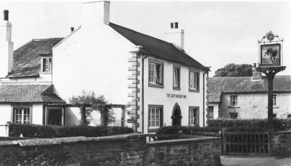 Old photograph of a whitewashed two-storey inn with stone quoins and The Greyhound Inn lettered on the front, a hanging sign of a running greyhound on a post, and a low stone wall in front
