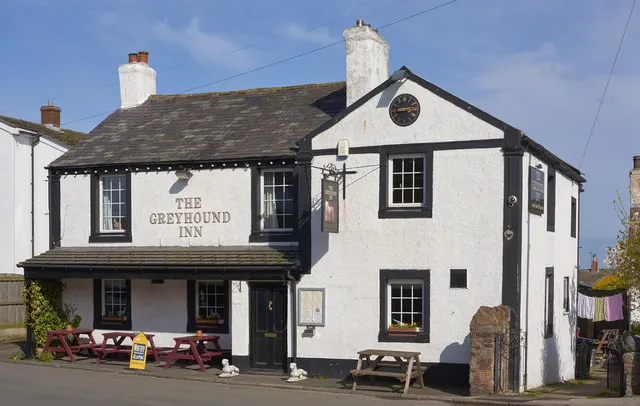Whitewashed two-storey village pub with a slate roof, black window surrounds and a clock on the gable, the words The Greyhound Inn painted in black on the front. A slate canopy runs over the ground floor, with red picnic benches and two small white dog statues by the door.