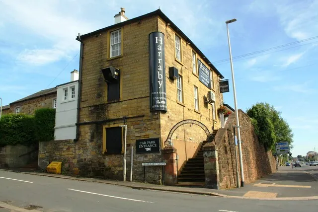 Tall yellow sandstone pub on a corner, three storeys with a slate roof and chimney, a long black vertical sign reading Harraby, Pub and Kitchen, Live Sports, Beer Garden, and stone steps under an iron arch rising beside a high stone wall, with a Car Park Entrance sign and Harraby Grove street sign below