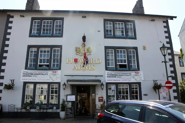 Three-storey white painted hotel front with dark window surrounds and black quoins, large gold letters reading Howard Arms beneath a gilded coat of arms and a Union flag. Banners for the Thwaites Hospitality Awards 2022 hang between the windows above a central doorway with lanterns, and a parked car cuts across the lower right corner.