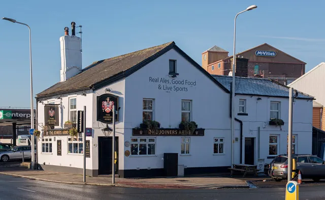Whitewashed pub with a dark slate roof and a tall white chimney, the gable end painted with Real Ales, Good Food and Live Sports, a hanging coat of arms sign and a long fascia reading The Joiners Arms, and a brick factory building with a McVitie's sign rising behind under a clear blue sky
