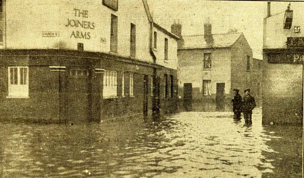 Newspaper photograph of a street under deep floodwater, with a corner pub lettered The Joiners Arms and a Church Street sign on the left, a row of houses beyond and two policemen standing in the water