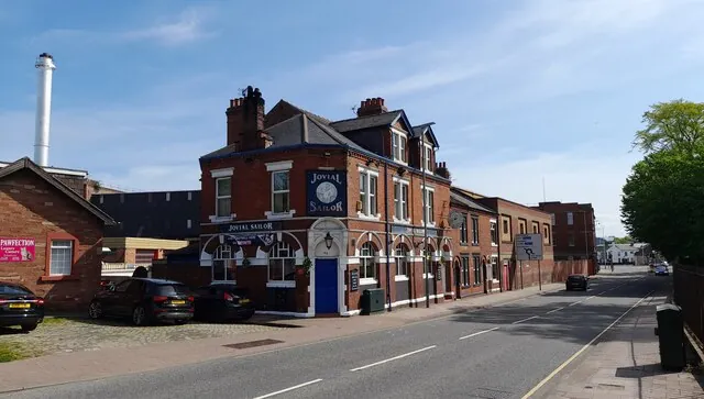 Red brick corner pub of two and three storeys with a slate roof and chimneys, white arched window heads on the ground floor, a blue door, and blue signs reading Jovial Sailor on the corner and above the door, beside a quiet road with a tall white chimney stack in the background