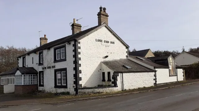 Whitewashed stone pub on a corner with a dark slate roof, brick chimneys and black quoins, the name Lane End Inn painted in black letters on both the gable end and the front. A white glazed porch sits at one end and lower whitewashed outbuildings run back along the road.