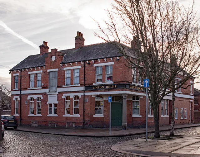 Large red brick corner pub of two storeys with a slate roof, tall chimneys and white painted window heads and string courses, a dark green door on the angled corner beneath a fascia reading Linton Holme, standing on a cobbled street with a bare tree in front
