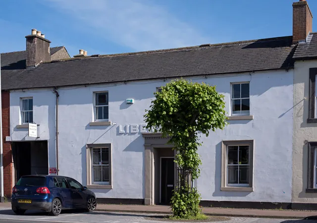Two-storey white rendered terrace building with a slate roof, a carriage arch at one end, and silver letters on the wall partly hidden behind a small clipped tree beside the doorway, with a dark car parked in front