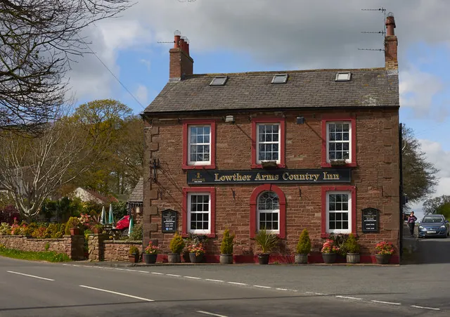 Two-storey red sandstone pub with a slate roof, end chimneys and white sash windows framed in red, a dark fascia reading Lowther Arms Country Inn above the door, and planters along the front